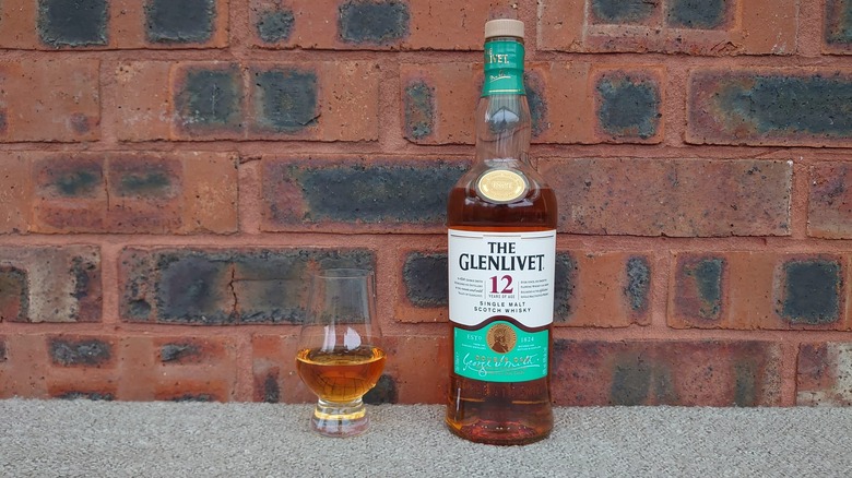 A bottle of The Glenlivet 12-year and a full glass posed in front of a brick wall