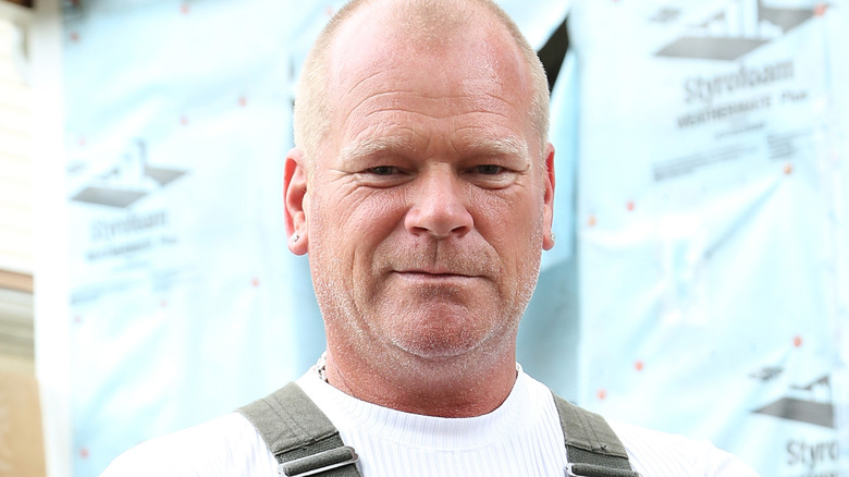 Mike Holmes in a white shirt and coveralls standing in front of a styrofoam insulation wall