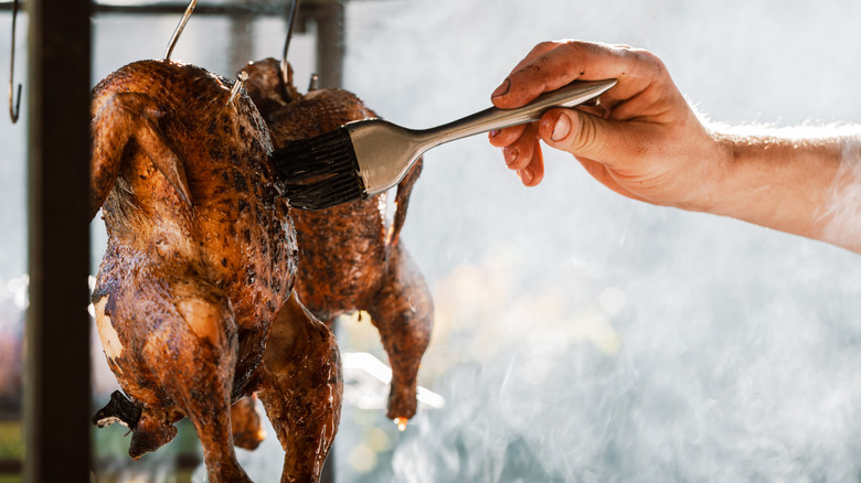 chef glazing a whole grilled chicken hanging on a hook