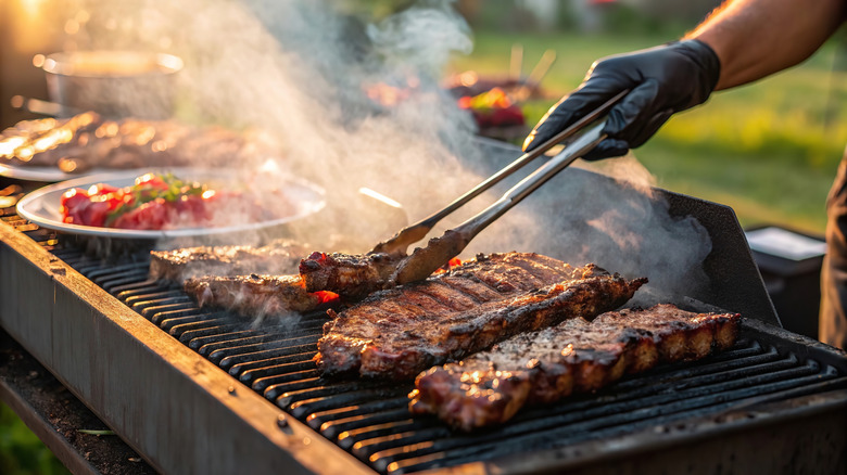 a hand with tongs picking up steak on a grill