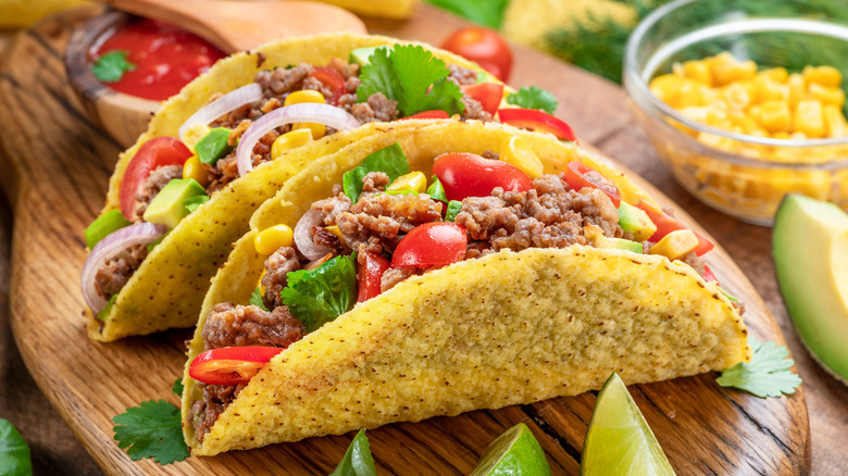 Two tacos loaded with meat and vegetables on a cutting board beside lime wedges and a bowl of corn