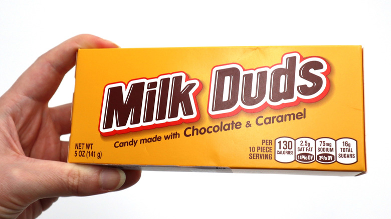 A hand holding up a yellow box of Milk Duds candy against a white background