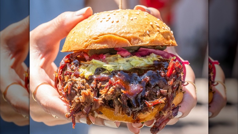 Hands holding a pulled pork sandwich with slaw and onions