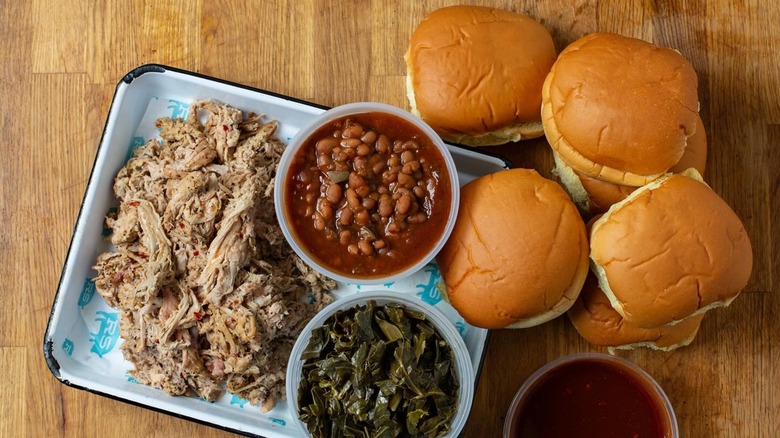 Pulled pork on a tray with beans, collard greens, buns, and BBQ sauce
