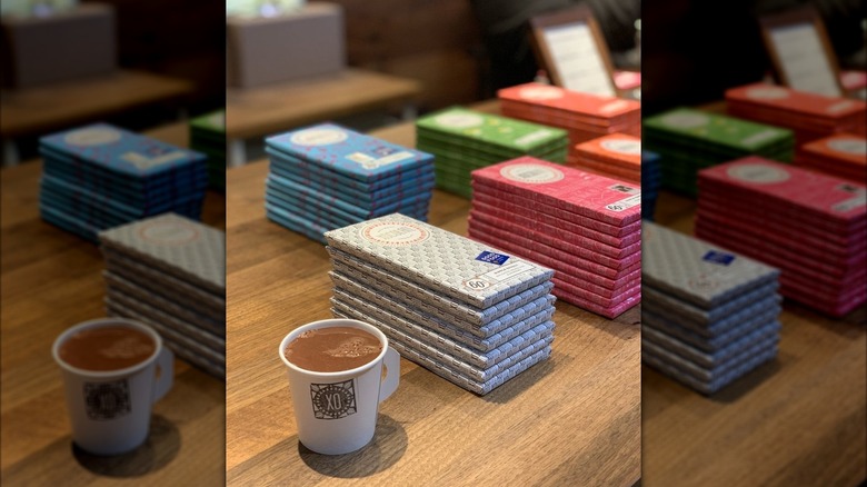 Small cup filled with drinking chocolate, on table alongside stacks of chocolate bars in various flavors