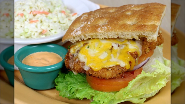 Fish sandwich on focaccia with cheese, tomato, onion, lettuce, next to sauce and with coleslaw in the background