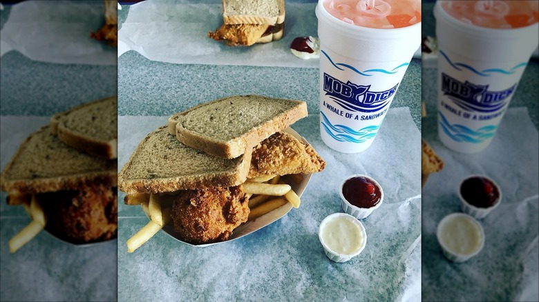 Fish sandwich next to soda and sauces on white table