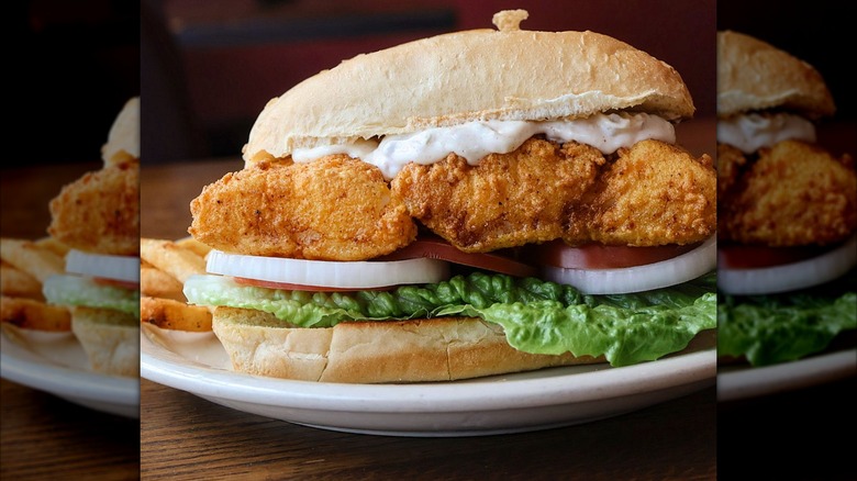 Fish sandwich with tartar sauce, lettuce, onion, tomato on white plate