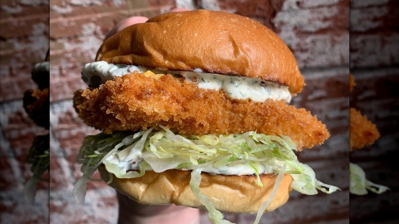 Close-up of hand holding a fish sandwich with tartar sauce and shredded lettuce.