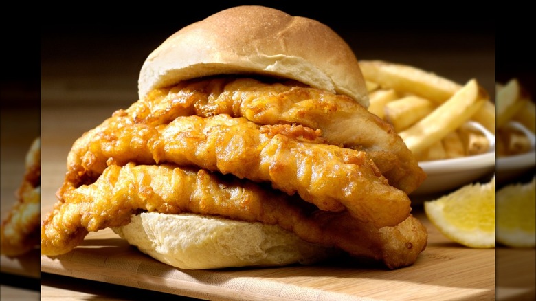 Fish sandwich in front of fries and lemon wedge on wooden cutting board