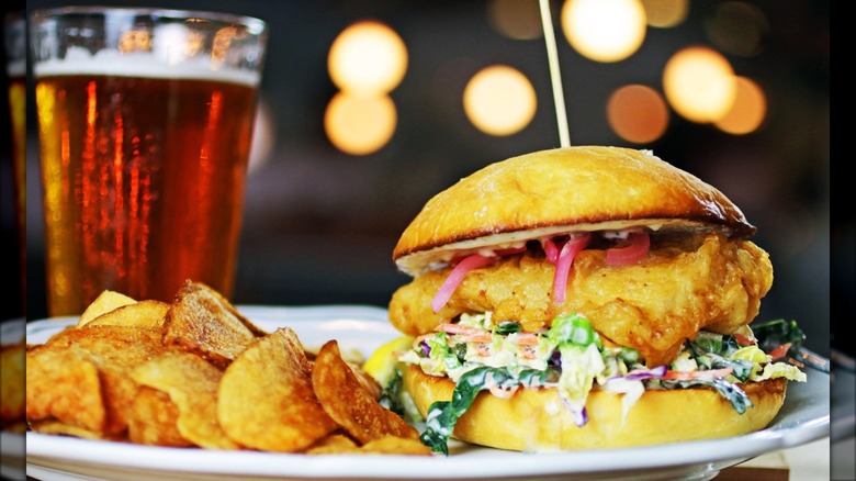 Fish sandwich next to potato chips and beer on white plate