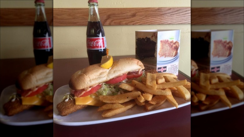 Fish sandwich with cheese, lettuce, tomato on hoagie roll with fries on white plate