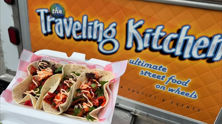 Three tacos in container in front of sign for Traveling Kitchen