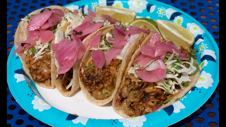 Four tacos on a paper plate with pickled veg and limes