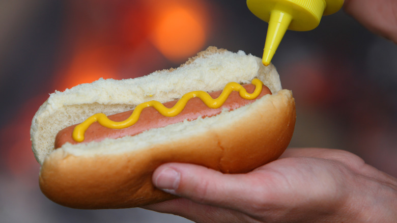 A hand holding a hot dog in a bun as mustard is applied to it