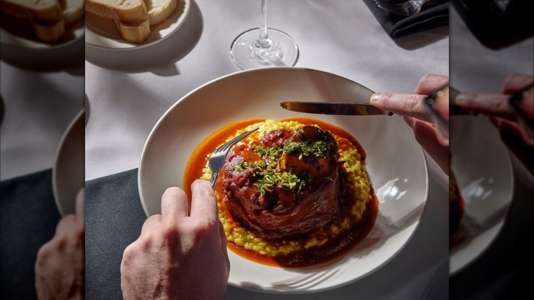 Osso bucco on white plate and elegant-looking table with hands, knife, and fork