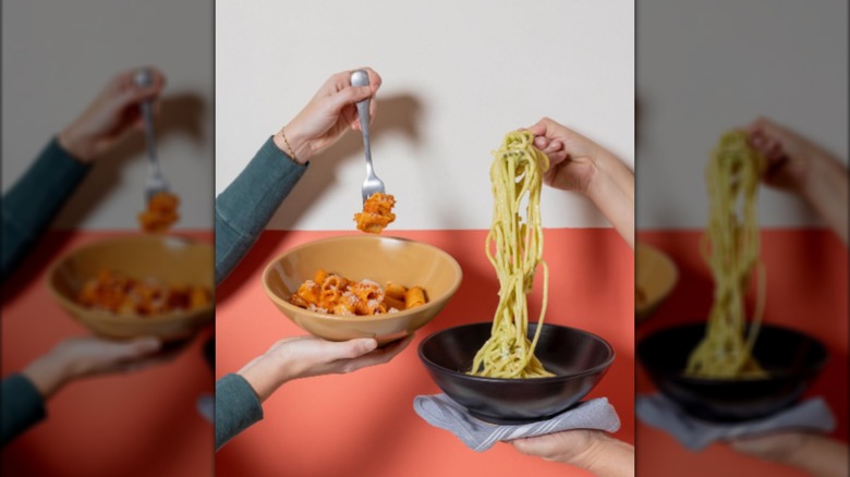 Hands holding bowls of pasta in front of red and white wall