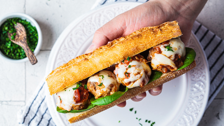 Hand holding a meatball sub over white plate on blue and white towel