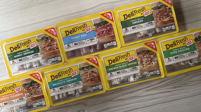 Multiple packages of the different kinds of Oscar Mayer brand Deli Fresh deli meats laid out uniformly on a light wood background