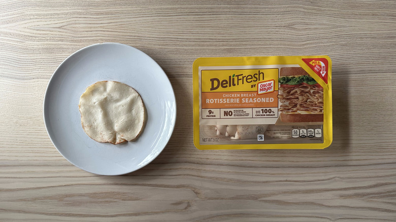 A package of Oscar Mayer Deli Fresh rotisserie seasoned chicken breast alongside a plate with a single slice of the chicken, both on a light wooden background