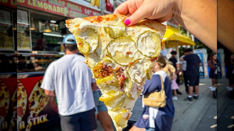Hand holding a slice of pickle pizza in front of fair food stands