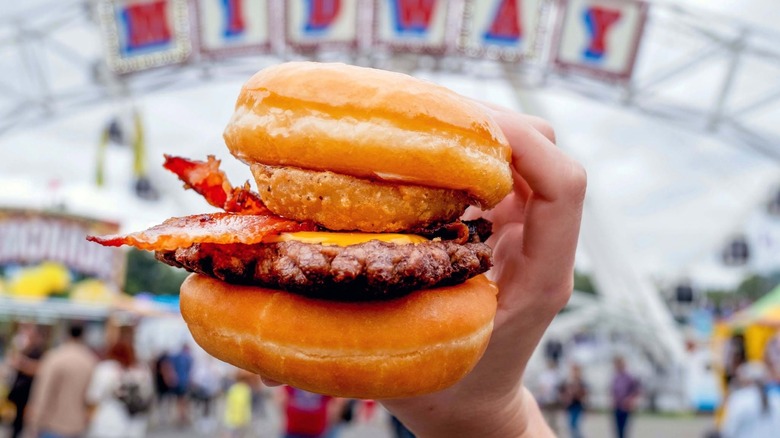 Hand holding donut burger with bacon in front of blurred midway