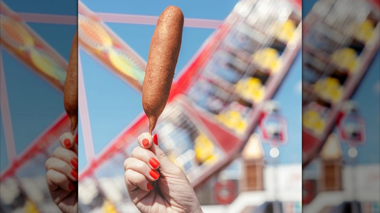 Hand holding a corn dog in front of blurred fair rides