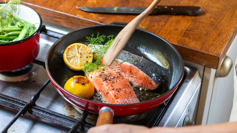 person cooking 2 salmon fillets in a pan on the stovetop, with lemon and herbs in the pan