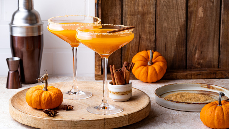 two autumnal orange-colored martinis on a tray beside cinnamon sticks and decorative pumpkins