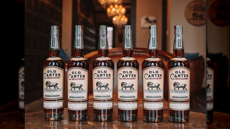 A row of 6 old carter bourbon bottles lined up on a wood table with a large leather chair and blurry parlor in the background