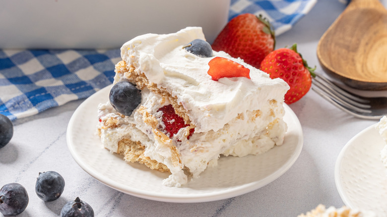 Square slice of cake with white cake layers, white whipped cream frosting, and strawberries and blueberries on round white plate surrounded by berries