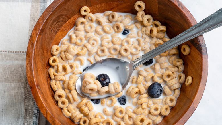 Honey Nut Cheerios are served in a wooden bowl with milk and blueberries.