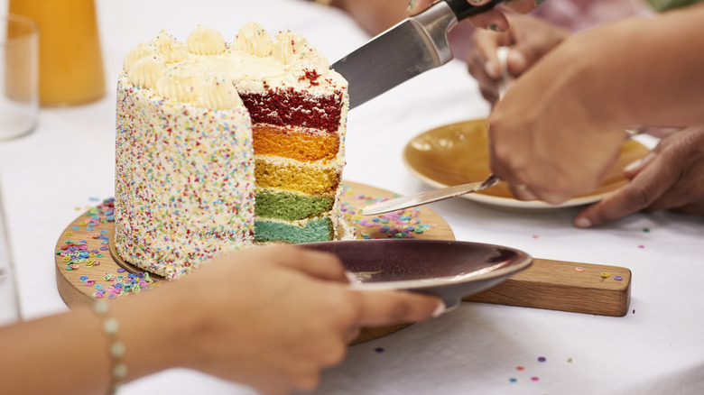 Hands slicing a small rainbow cake and perparing to serving it to party guests