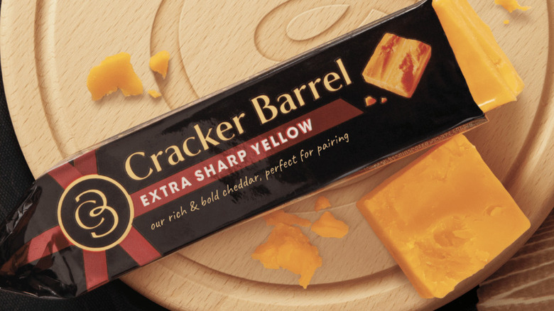 A block of Cracker Barrel extra sharp yellow cheddar cheese on a wooden cheese board with a few slices and chunks broken off