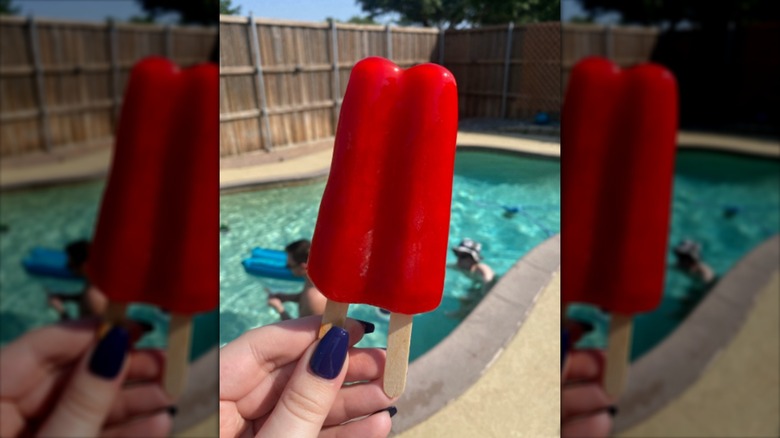 Someone with blue nail polish holding a red Twin Pop with a pool in the background