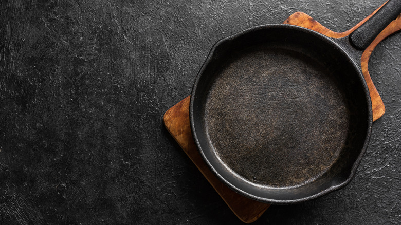 A cast iron pan on a wooden heating pad sitting on top of dark textured surface.
