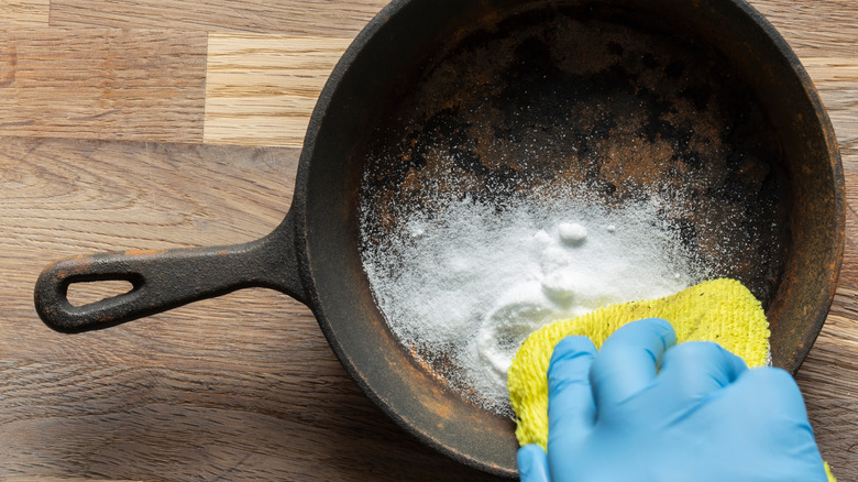 Someone wearing blue gloves and using a yellow cloth to scrub a rusty cast iron pan with baking soda