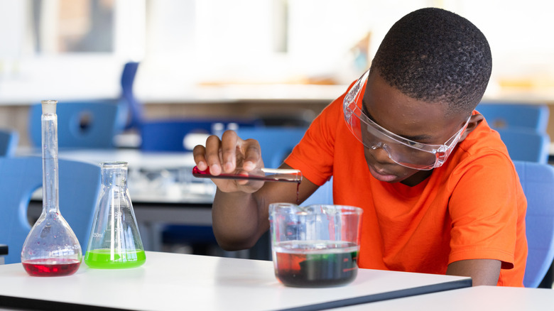 a child wearing safety glasses pouring dark liquid from a test tube into a larger beaker in a classroom setting