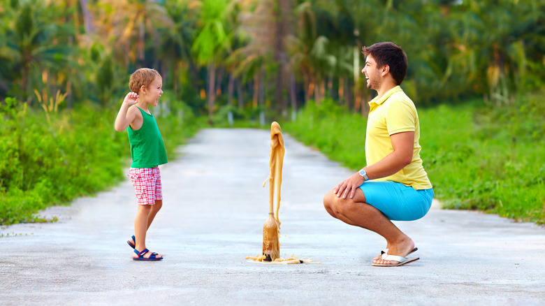 a father and his young son smile at each other on an outdoor path as a geyser of soda shoots out of the top of a plastic bottle
