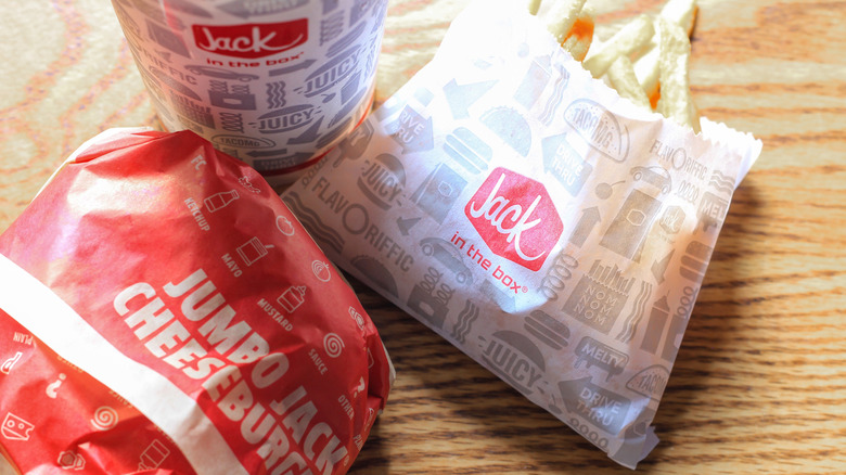 A wrapped Jack in the Box cheeseburger, sitting on a wooden table next to a bag of Jack in the Box fries and a Jack in the Box branded drink