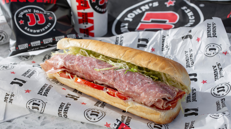 A Jimmy John's sub sandwich on top of wrapper next to bag of chips and drink