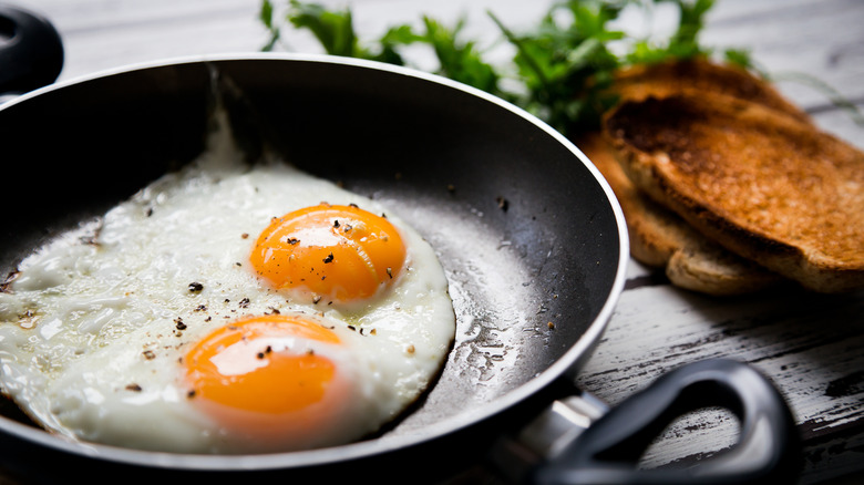 two sunny-side up fried eggs in a skillet, tossed with black pepper. There is toast and some greens in the background