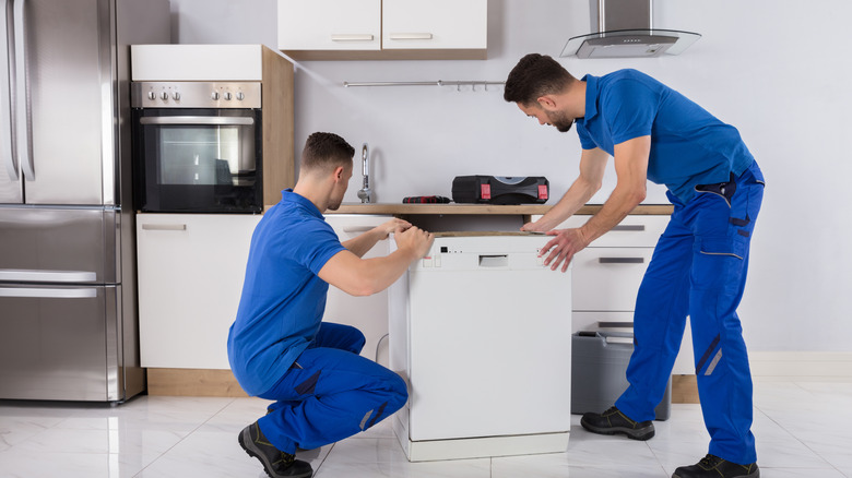 Two male workers wearing blue jumpsuits moving a dishwasher in a kitchen.