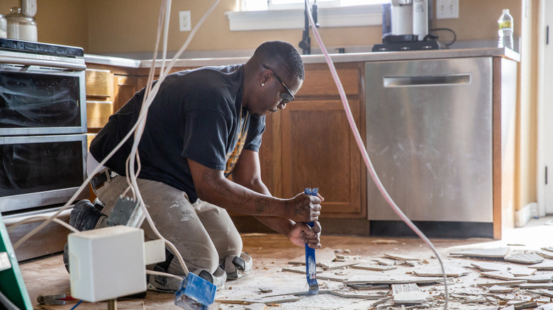 A man in overalls and safety glasses removing tile flooring in a kitchen during a renovation