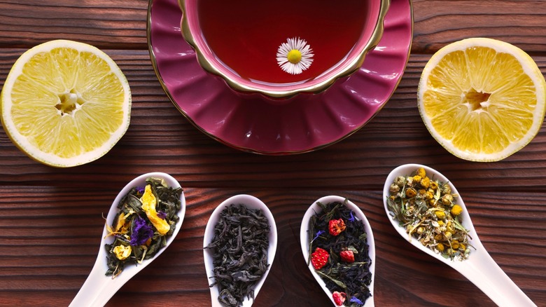 Several spoons or different loose leaf teas and a pair of lemon halves around a teacup