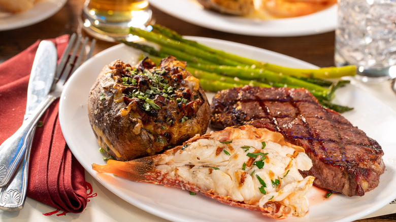 A grilled steak on a plate with a lobster tail, baked potato, and asparagus
