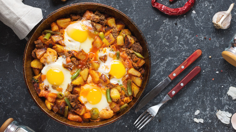 Skillet of breakfast hash with potatoes, vegetables, and fried eggs next to cutlery and ingredients