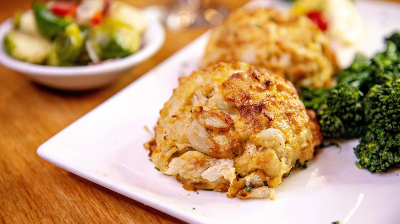 A white plate of crab cakes and broccoli on a restaurant table next to a bowl of brussels sprouts