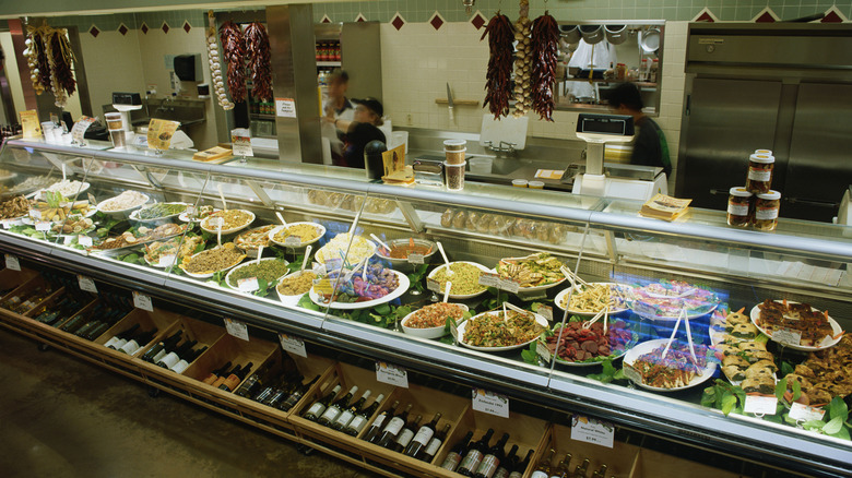 A refrigerated deli counter, filled with platters of various deli dishes, with several employees behind the counter