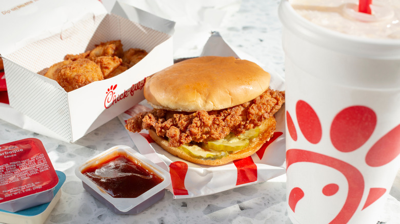 A Chick-fil-A sandwich sits next to nuggets, an open sauce packet, and a drink.